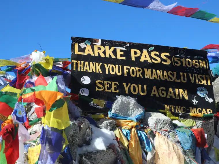 Prayer flags around the sign on the pass, which reads Larke Pass 5106m, thank you for Manaslu visit, see you again