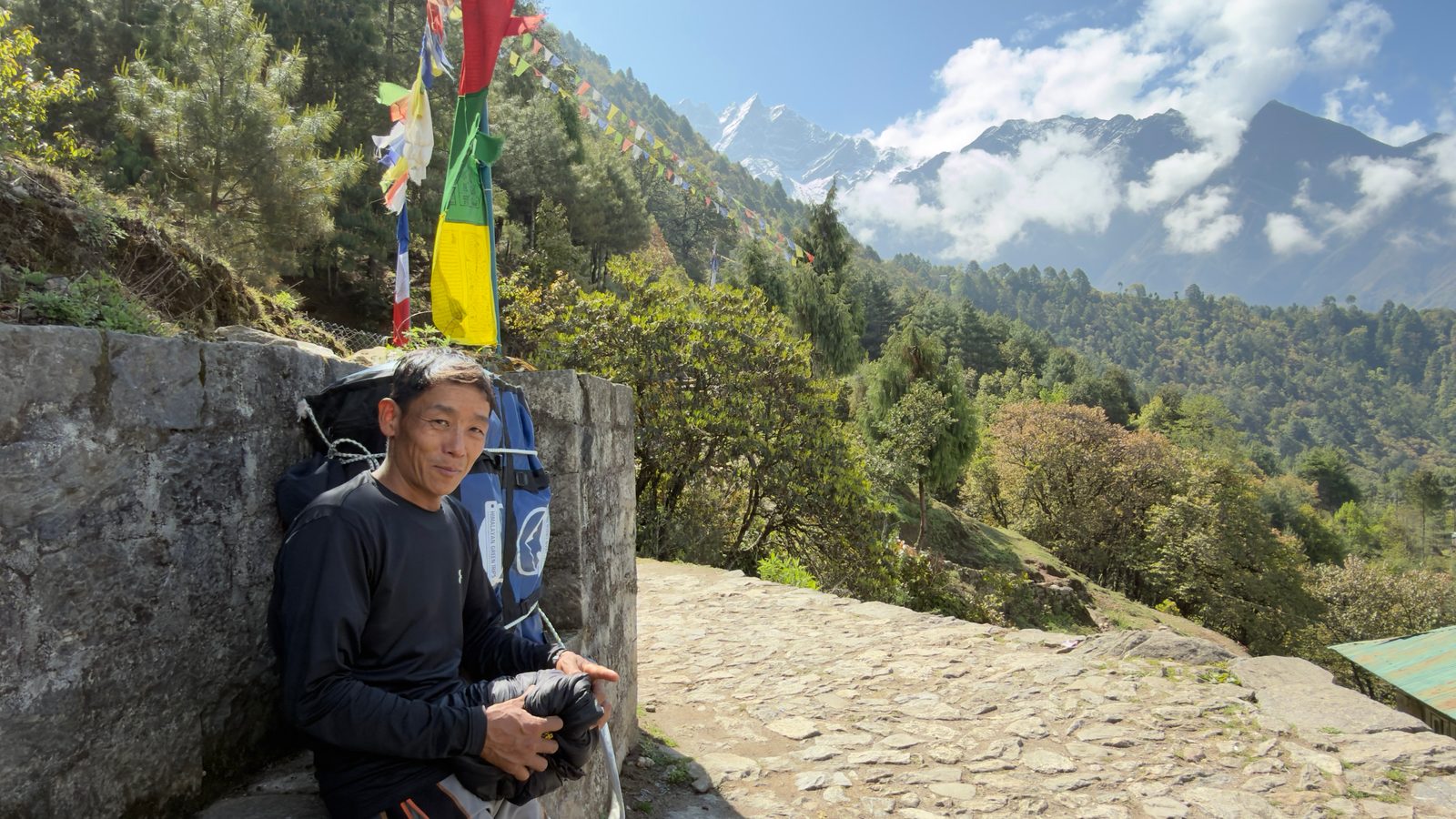 A porter resting beside his backpack and a stone wall, with prayer flags and mountains behind